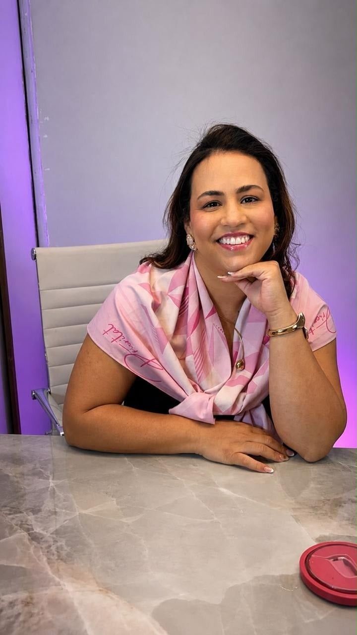 Micaele Amorim, com lenço rosa, sorrindo apoiada na mesa da clínica