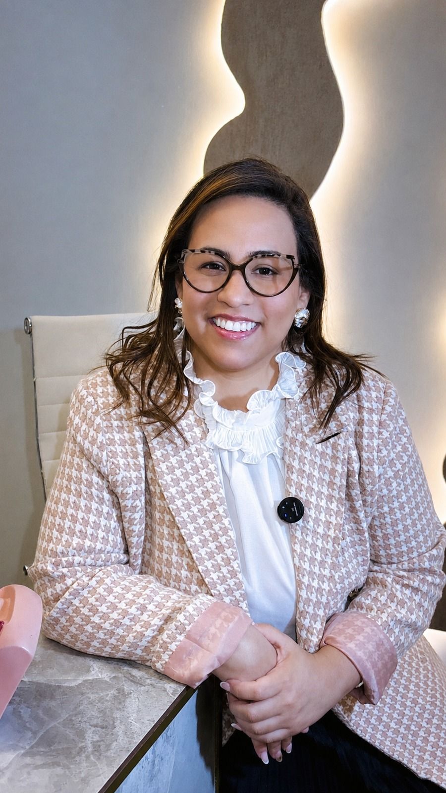 Micaele Amorim, de blazer, sorrindo diante do painel iluminado do consultório